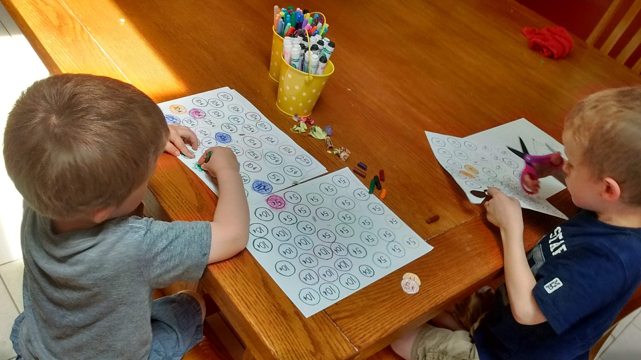 boys coloring paper coins with tiny broken crayons and cutting them out with scissors