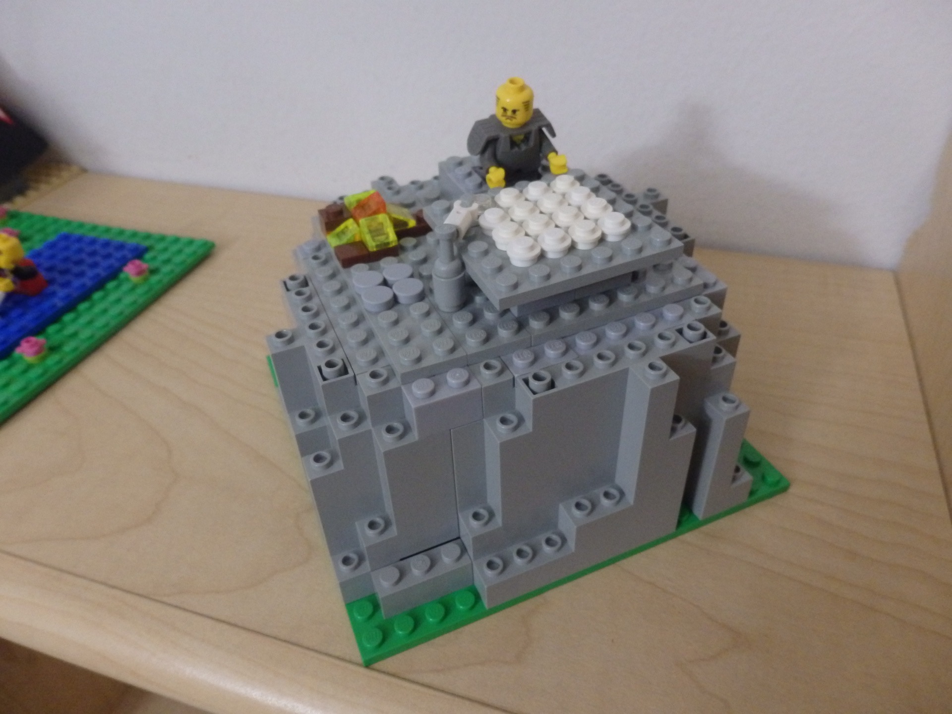 lego mountain with a flat top, man kneeling in front of an alter with 15 white round lego pieces, and 1 clear, with a white lego hand reaching for the clear stone