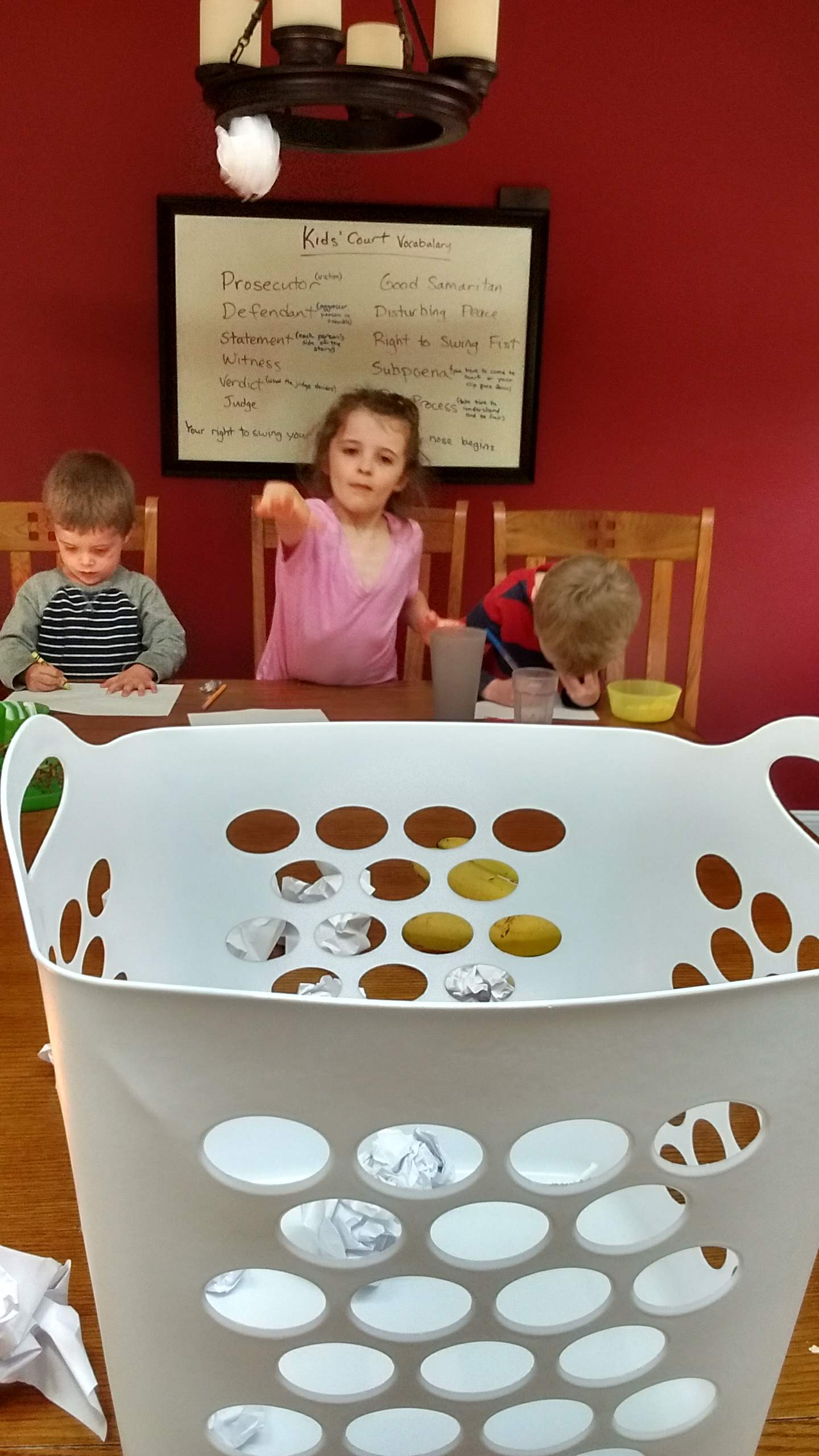 three kids writing on paper, crumpling them and throwing them in the basket