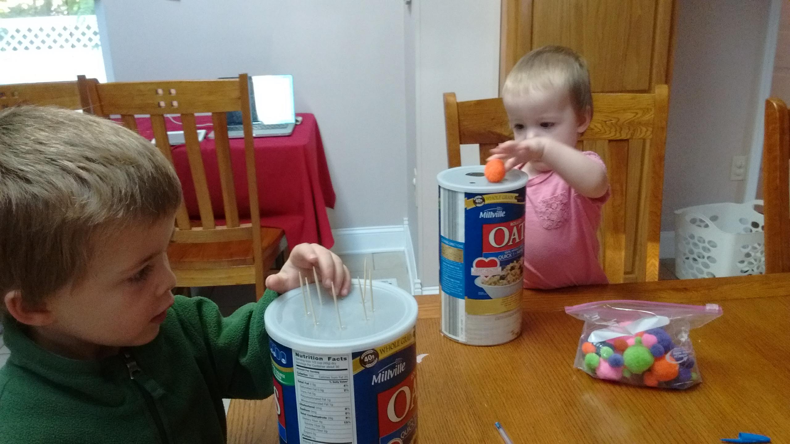 preschooler putting toothpicks in oats jar with pinholes cut and a baby putting pom pom in holes